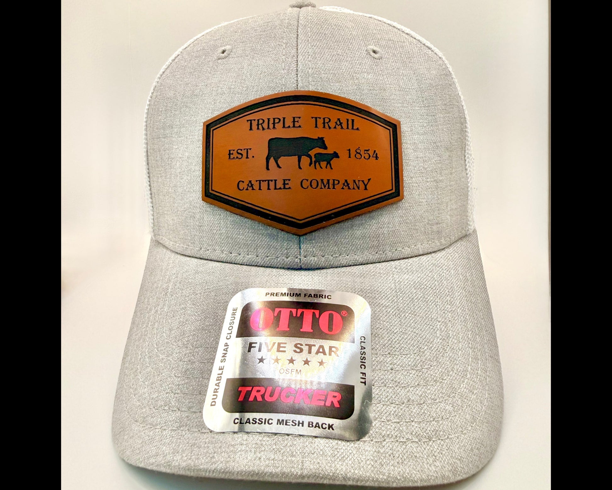 A light gray heather trucker cap with a brown leather patch on the front reading 'TRIPLE TRAIL CATTLE COMPANY EST. 1854'.