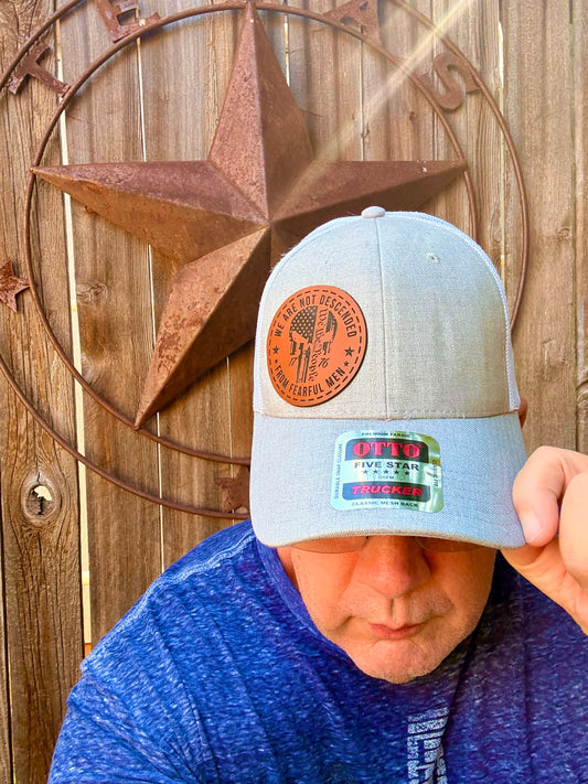 A man wearing a light gray trucker hat with a brown circular patch on the front and a blue heathered t-shirt, posing in front of a rustic wooden wall with a large metal star.