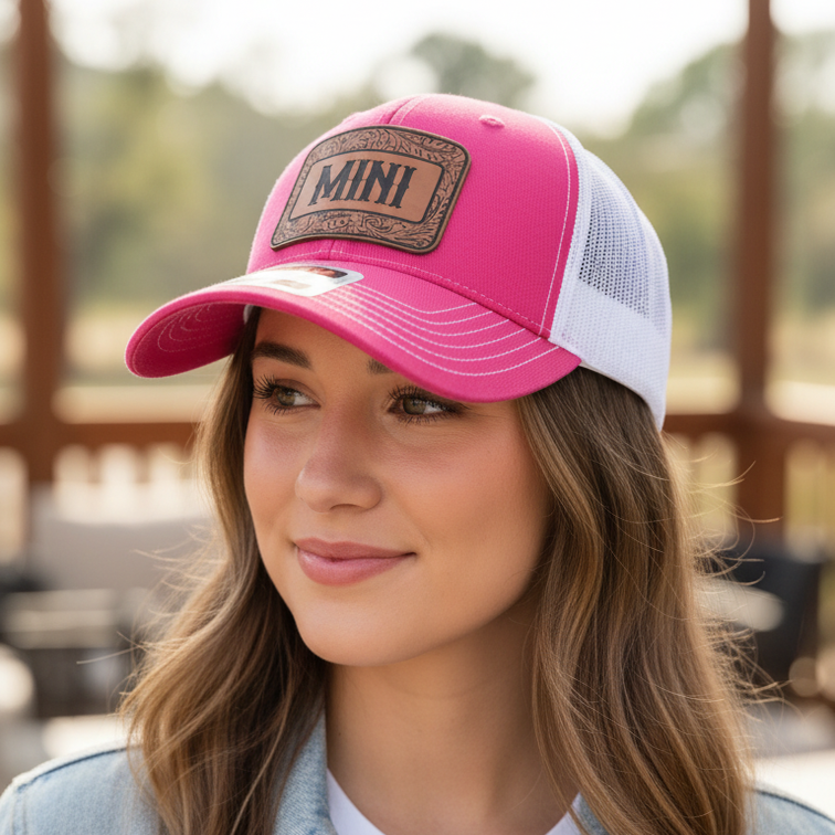 A woman wearing a pink and white trucker hat with a brown 'MINI' patch on the front, styled with a light blue denim jacket over a white t-shirt in an outdoor setting.