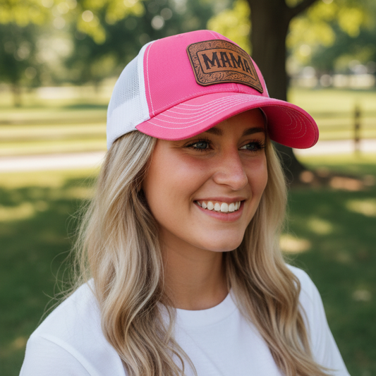 A woman outdoors wearing a pink and white trucker hat with a brown leather patch that reads 'MAMA'.