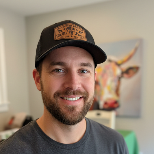 A smiling man wearing a black baseball cap with a brown leather patch that reads 'OCEAN WATERS' and a dark gray t-shirt, standing in a home interior with a cow painting on the wall behind him.