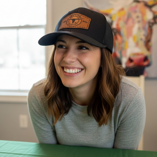 A smiling woman with shoulder-length brown hair wearing a black baseball cap with a brown leather patch logo, sitting behind a green table in an indoor setting.