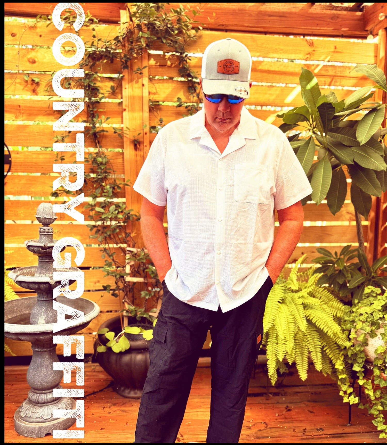 A man wearing a white short-sleeve button-up shirt, dark pants, a white cap, and blue sunglasses stands on a wooden deck in front of a wooden slat wall with plants. The text 'COUNTRY CRAFTED' is vertically written on the left side.